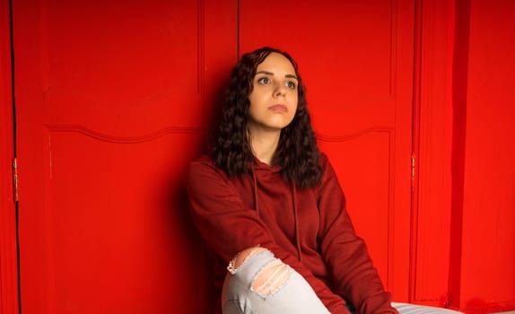 Young woman sitting on floor. Curly brunette poses near red wall