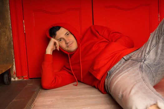 Young handsome male in hood lying on floor. Thoughtful man poses near red wall