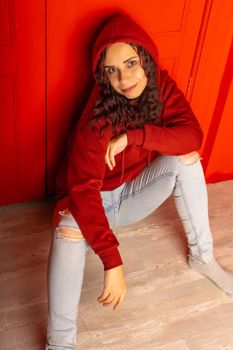 Young woman in hood sitting on floor. Curly brunette poses near red wall