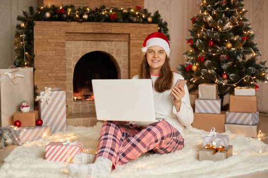 Smiling young woman with laptop sitting near Christmas tree and present boxes, spending New Year eve working online, holds smart phone in hands, wearing checkered pants, shirt and santa claus hat.