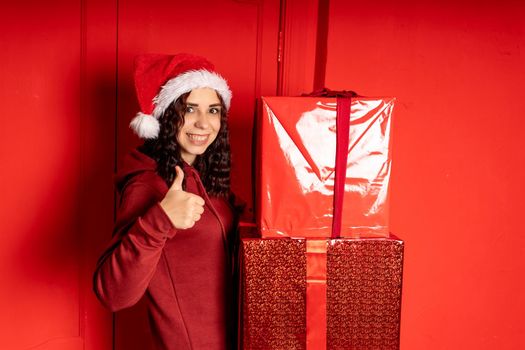 Young woman in Santa Claus hat holds big gifts, standing near red wall. Happy female in Christmas hat with boxes of gift. Concept of holidays, presents and good mood