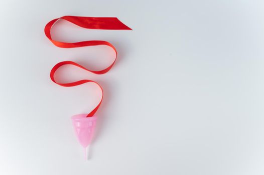 Pink menstrual cup and red satin ribbon on a white background. Copy space.