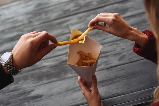 One french fries for two. Young couple are eating french fries at the street food market. Cold season. Close-up photo of couple eating from one package