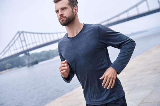 Morning workout. Close up of young man in sports clothing exercising while jogging on the bridge outdoors