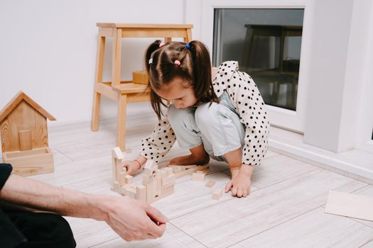 Girl sits on the floor in a room and plays with her dad in a natral wood constructor. Wooden childrens construction kit. Toys made of eco-friendly material. The castle is made of wooden cubes.