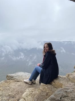 Young woman sitting on edge of cliff in foggy and cloudy weather. Female tourist enjoys spectacular view of nature, sitting on high rock