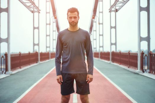 Confident sportsman. Portrait of young and handsome bearded man in sports clothing standing on the bridge and looking at camera. Sport concept. Healthy lifestyle