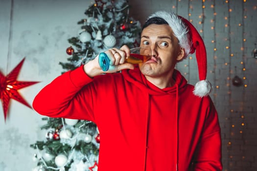 Young man in santa hat with glass of champagne on background of Christmas tree. Adult male posing at coniferous tree with decorative adornments. Concept of Christmas celebration at home