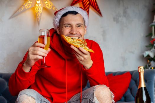 Young man in santa hat drinks champagne and eats pizza, sitting on sofa in room. Happy male resting with food and alcohol. Concept of Christmas celebration at home