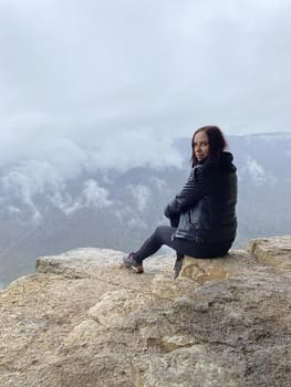 Young woman sitting on edge of cliff in foggy and cloudy weather. Female tourist enjoys spectacular view of nature, sitting on high rock