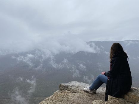 Young woman sitting on edge of cliff in foggy and cloudy weather. Female tourist enjoys spectacular view of nature, sitting on high rock
