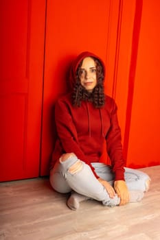 Young woman in hood sitting on floor. Curly brunette poses near red wall
