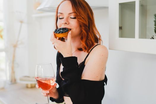 The girl is sitting eating a cake and drinking wine. A young girl in a black negligee is sitting in the kitchen eating a cake and drinking wine. Bright kitchen. Sexy girl is resting.