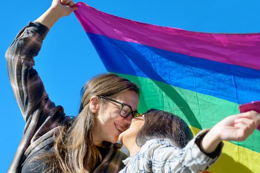 Affectionate lesbian couple holding an LGBT flag under blue sky. High quality photo.