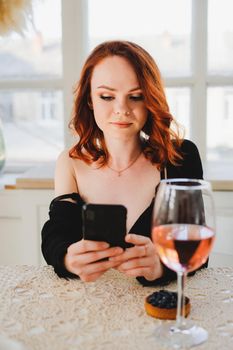 A beautiful girl with red hair holding a smartphone. There's a glass of rose wine on the table. The girl is resting.