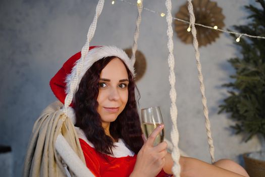 Young woman in Santa Claus suit with glass of champagne sitting on hammock. Attractive female in Christmas hat swinging on hammock chair. Concept of Christmas celebration at home