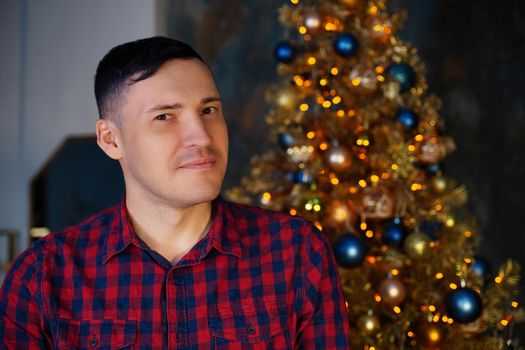 Young handsome man in plaid shirt on background of Christmas tree. Adult attractive male posing at coniferous tree with decorative adornments. Concept of Christmas celebration at home