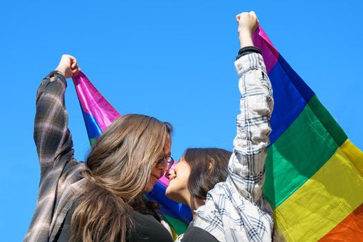Affectionate lesbian couple holding an LGBT flag under blue sky. High quality photo.