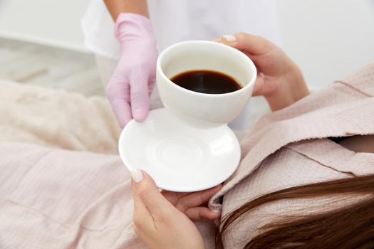 Beautician serving cup of coffee to her client after body treatment in beauty salon. Relaxing with beverages on cosmetic bed. Beauty , pleasure concept