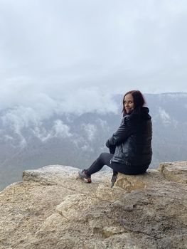 Young woman sitting on edge of cliff in foggy and cloudy weather. Female tourist enjoys spectacular view of nature, sitting on high rock