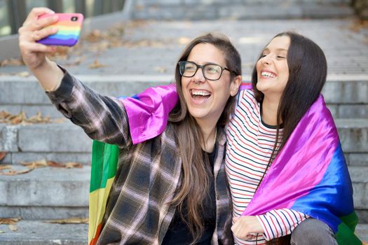 Cheerful happy lesbian couple holding lgbt flag taking a selfie outdoors. High quality photo