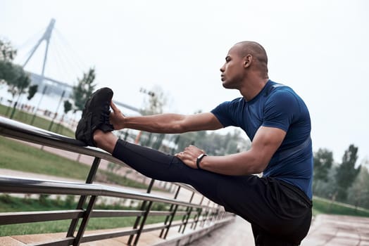 A close-up portrait of an african fitness man stretching legs on a national park background. Side view