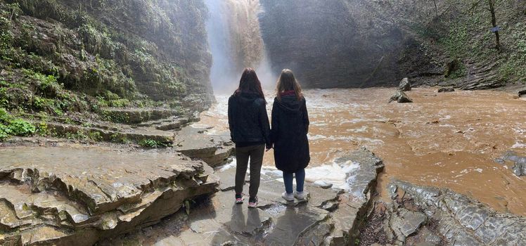 Rear view of two women looking on powerful muddy waterfall. Tourists standing on rocks in mountainous terrain and enjoying beautiful view of cataract