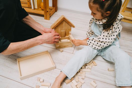 A girl and her father play a wooden constructor. Build a tower of wooden cubes.