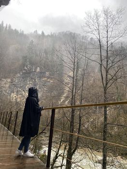 Side view of young woman standing on platform in mountainous terrain. Female tourist enjoys spectacular view of mountain landscape in rainy and cloudy weather