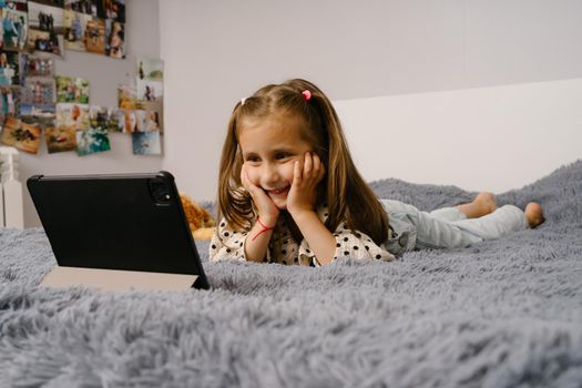 The girl lies on the bed with a gray fluffy blanket and looks at the computer tablet. Little girl laughs and watches cartoons.