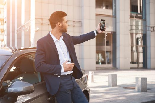 Visiting car dealership. Handsome man is doing selfie with his new car, showing thumb and smiling