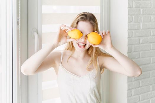 Photo of a beautiful woman covering eyes with citrus lemons posing by the window in a white room