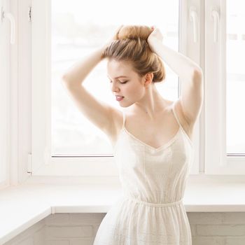 beautiful young woman in a light white dress posing by a large window