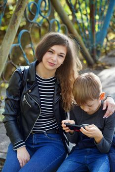 A focused boy is playing a game on a smartphone in the park. A serious focused school age boy in casual clothes is sitting on a bench and together with his mother and playing a game on a mobile phone.