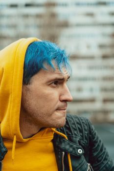Portrait of young man on background of high-rise building. Handsome guy with blue hair posing on city street in springtime.