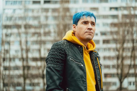 Portrait of young man on background of high-rise building. Handsome guy with blue hair posing on city street in springtime.