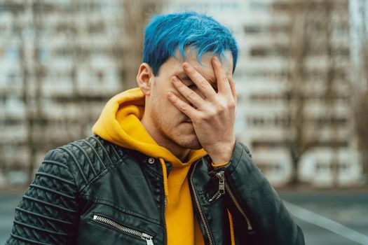 Portrait of young man closing his face with hand on background of high-rise building. Handsome guy with blue hair posing on city street in springtime