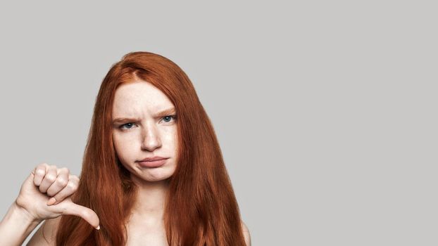 It's not funny. Studio shot of redhead girl showing thumb down and making upset face while standing against grey background. Negative emotions