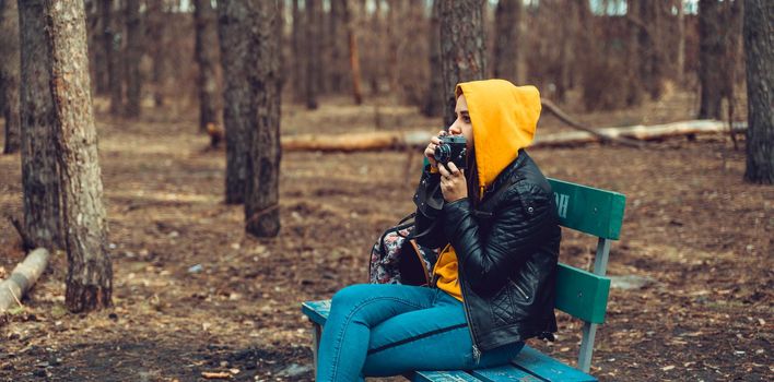 Young woman in casual clothes sitting on bench and photographing on old photo camera in forest. Female resting and taking photos with old camera in early spring