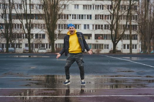 Young man standing in puddle on sports stadium on background of high-rise building. Happy guy with blue hair having fun on city street in springtime