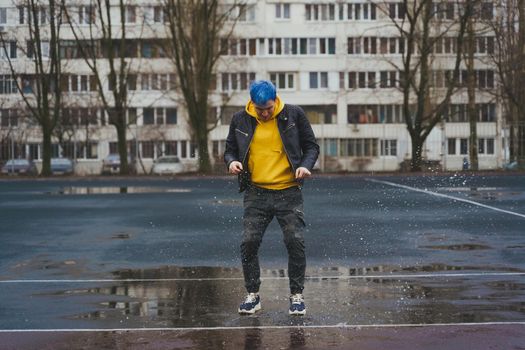 Young man jumps in puddle on sports stadium on background of high-rise building. Happy guy with blue hair having fun on city street in springtime
