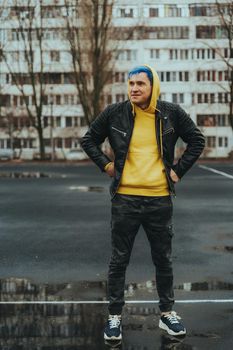 Young man standing on sports stadium on background of high-rise building. Handsome guy with blue hair posing on city street in springtime