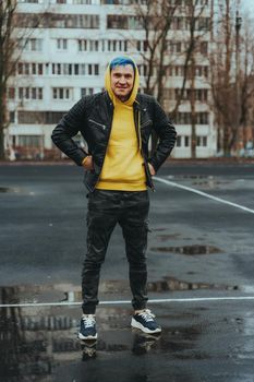 Young man standing on sports stadium on background of high-rise building. Handsome guy with blue hair posing on city street in springtime