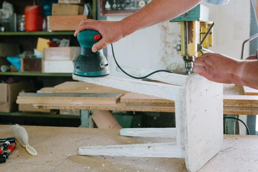 A man is polishing a white old stool. Grinding machine. Restoration of furniture.