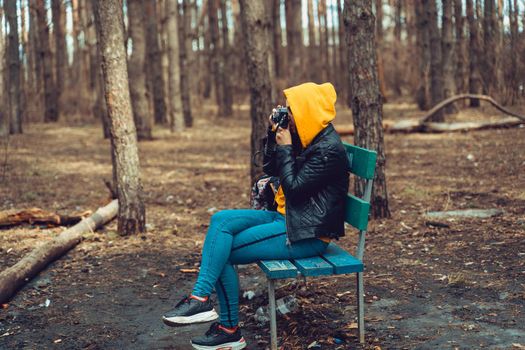 Young woman in casual clothes sitting on bench and photographing on old photo camera in forest. Female resting and taking photos with old camera in early spring