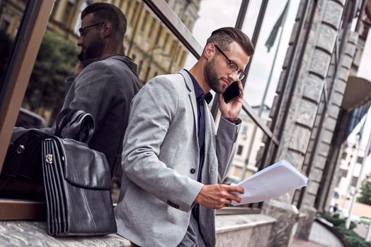 Business communications. Young businessman standing leaning on wall on the city street talking on smartphone with partner reading documents