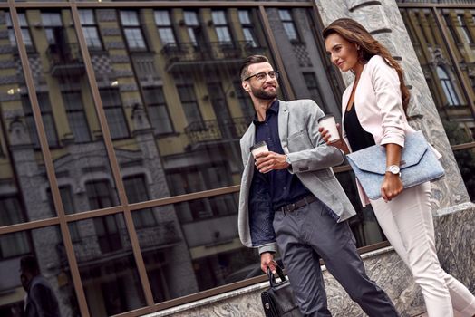 Hurry for meeting. Two young business people walking outside on the city street drinking hot coffee talking smiling joyful close-up