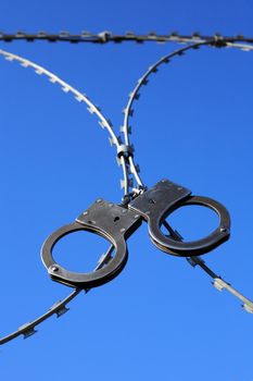 Old metal handcuffs hanging on razor wire against blue sky