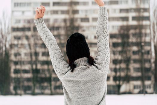 Rear view of young woman in gray knitted sweater and hat standing on street in winter season. Female looking at high-rise building and stretching her hands in cloudy weather