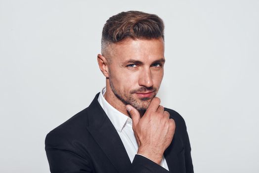 Studio shot of handsome and confident bearded businessman in classic suit touching his chin while standing against grey background. Studio shot. Business. Men beauty